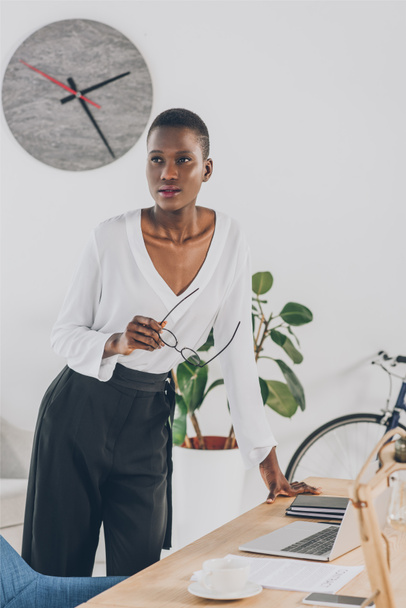 fashionable attractive african american businesswoman holding glasses and leaning on table in office - Photo, Image