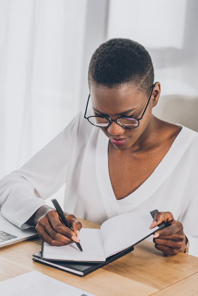 elegante atraente mulher de negócios afro-americana escrevendo algo para notebook no escritório
 - Foto, Imagem