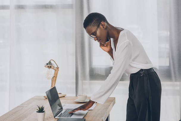side view of stylish attractive african american businesswoman talking by smartphone and using laptop in office - Photo, Image