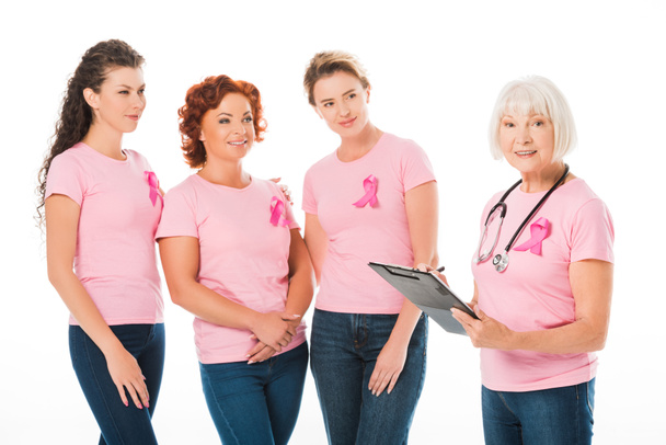 женщины в розовых футболках с лентами для информирования о раке молочной железы смотрят на улыбающегося старшего врача со стетоскопом и блокнотом, изолированными на белом
 - Фото, изображение
