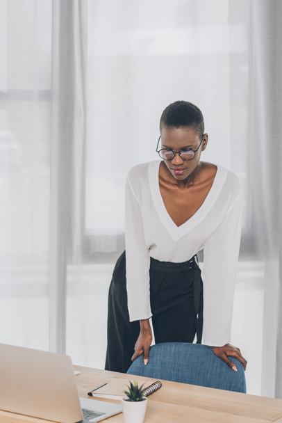 Stylová atraktivní africká americká podnikatelka opírající se o křeslo a při pohledu na přenosný počítač v kanceláři - Fotografie, Obrázek