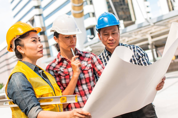 Three industrial engineer wear safety helmet engineering working and talking with drawings inspection on building outside. Engineering tools and construction concept. - 写真・画像