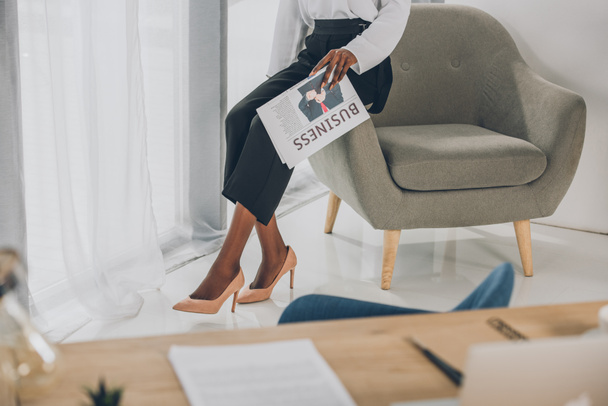 imagen recortada de la mujer de negocios afroamericana sentada en un sillón y sosteniendo el periódico en la oficina
 - Foto, Imagen