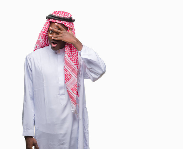 Αραβικά αφρικανικά νεαρός φορώντας παραδοσιακές keffiyeh πέρα από το απομονωμένο υπόβαθρο peeking σε κατάσταση σοκ που καλύπτουν το πρόσωπο και τα μάτια με το χέρι, κοιτάζοντας μέσα από τα δάχτυλα με αμηχανία έκφρασης. - Φωτογραφία, εικόνα