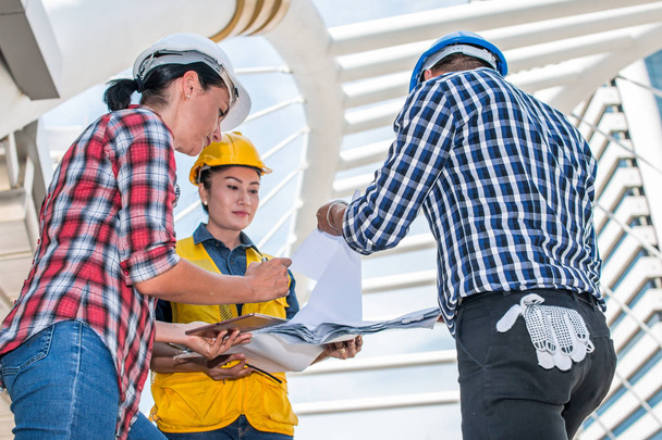 Three industrial engineer wear safety helmet engineering working and talking with drawings inspection on building outside. Engineering tools and construction concept. With copy space - Photo, Image