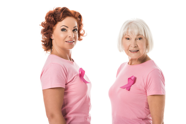 vista lateral de las mujeres con cintas de color rosa sonriendo a la cámara aislada en blanco, concepto de conciencia de cáncer de mama
 - Foto, Imagen