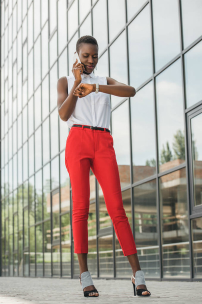 стильный привлекательный африканский американский бизнесмен говорит по смартфону и проверяет время рядом с бизнес-центром
 - Фото, изображение