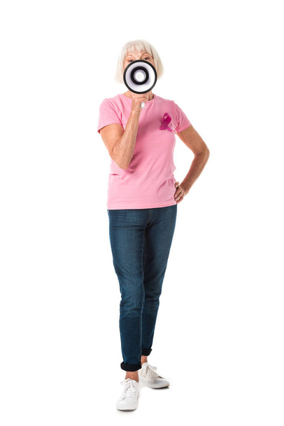 Ganzkörperansicht der Seniorin in rosa T-Shirt mit Brustkrebs-Bewusstseinsband, Megafon in der Hand und Blick in die Kamera isoliert auf weiß - Foto, Bild