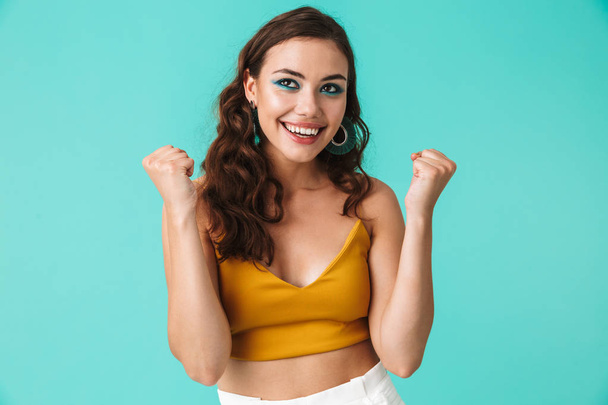 Photo of adorable charming girl 20s with beautiful makeup rejoicing and clenching fists in happiness isolated over blue background - Photo, Image