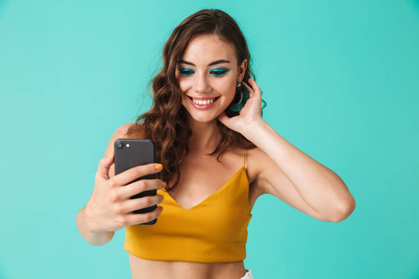 Image of glamorous european woman 20s with beautiful makeup smiling while holding smartphone and taking selfie isolated over blue background - Fotó, kép
