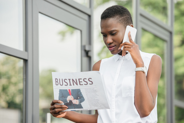 Stylová atraktivní africká americká podnikatelka mluví o smartphone a čtení noviny na ulici - Fotografie, Obrázek