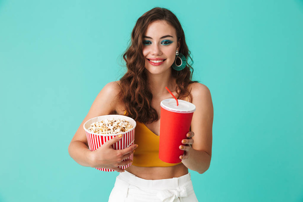 Photo of charming young woman 20s wearing fashion earrings holding bucket with popcorn and drinking soda from big red paper cup isolated over blue background - Φωτογραφία, εικόνα