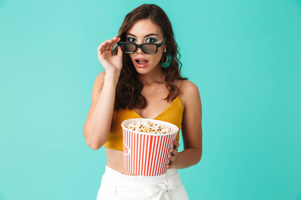 Photo of fashionable excited woman 20s wearing glasses holding bucket with popcorn and looking at camera isolated over blue background - Foto, afbeelding