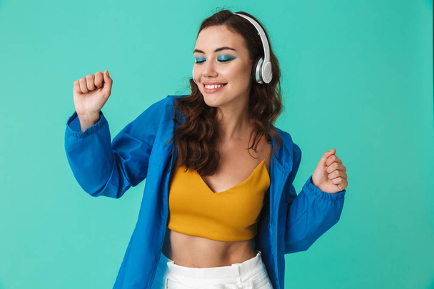 Image of caucasian brunette woman 20s wearing raincoat or jacket dancing while listening to music via wireless earphones isolated over blue background - Fotoğraf, Görsel