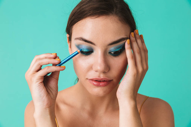 Close up portrait of a pretty young girl with bright make-up holding blue eye pencil isolated over blue - Foto, Bild