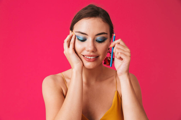 Image of lovely posh woman 20s wearing earrings smiling and applying makeup with blue eyeliner isolated over pink background - Valokuva, kuva