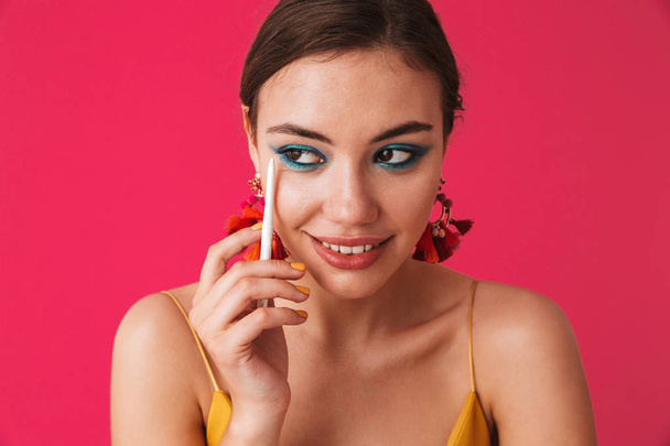 スタイリッシュなトレンディな女性 20 代着用イヤリングよそ見とピンク背景に分離された化粧用白鉛筆を保持の写真 - 写真・画像