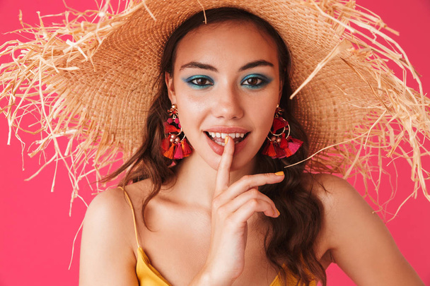 Close up of a beautiful young girl with bright make-up with earrings and summer hat posing isolated over pink - Zdjęcie, obraz