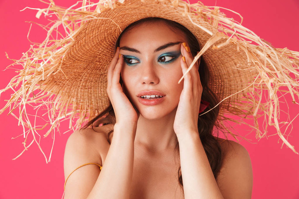 Image closeup of trendy fashionable woman 20s wearing big straw hat holding face and looking aside isolated over pink background - Фото, зображення