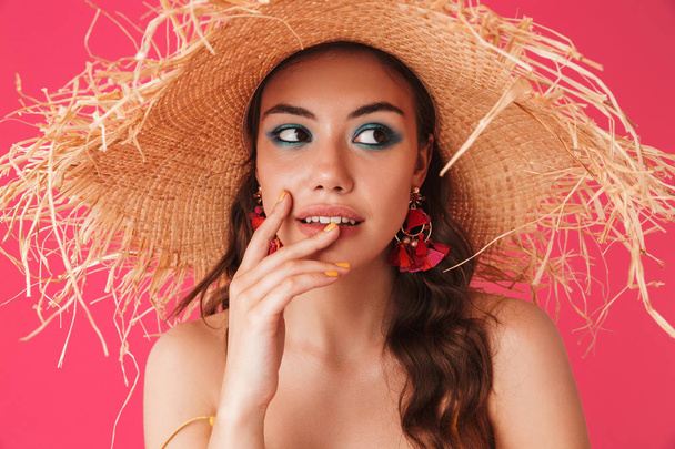 Close up of a pretty young girl with bright make-up with earrings and summer hat posing isolated over pink - Zdjęcie, obraz