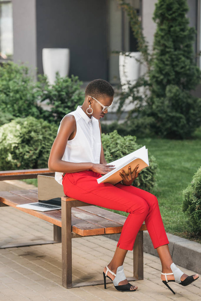 fashionable african american businesswoman sitting on bench and reading documents on street - Фото, изображение