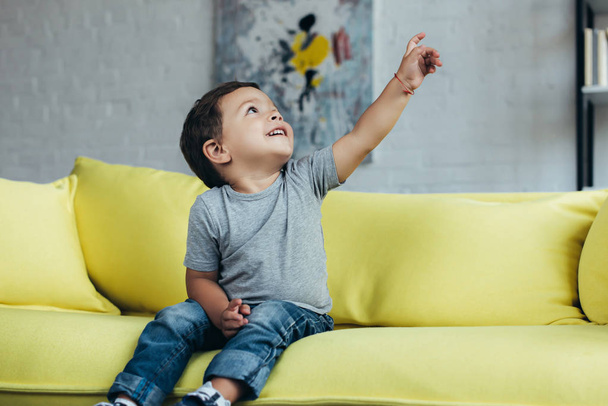 usměvavý chlapeček směřující nahoru, zatímco sedí na žluté pohovce doma - Fotografie, Obrázek