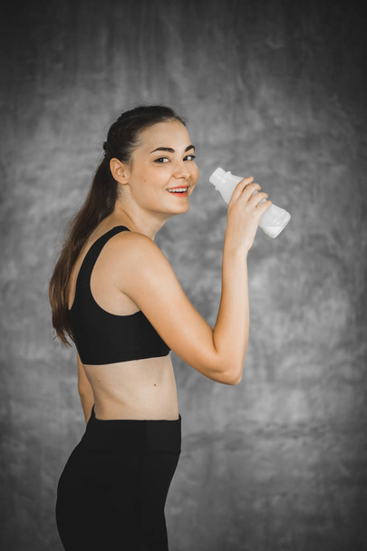 Młoda kobieta picia mleka z butelki po wysiłku w siłowni sprawny. Piękne kobiety na siłowni przerwę z piciem zdrowie i uśmiech w sportowej. Pojęcie zdrowego, fitness, sport, styl życia. - Zdjęcie, obraz