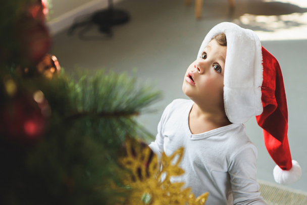 adorable little kid in santa hat looking up at christmas tree - Photo, Image