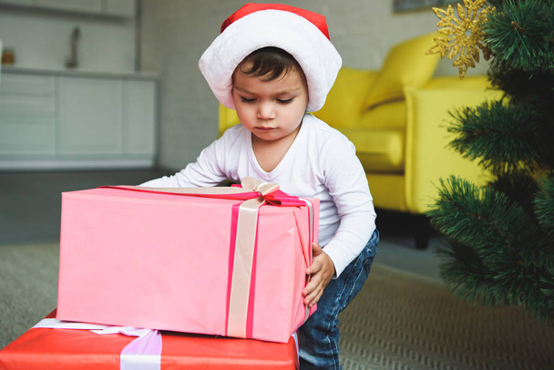 adorable boy in santa hat with gift boxes near christmas tree - Zdjęcie, obraz