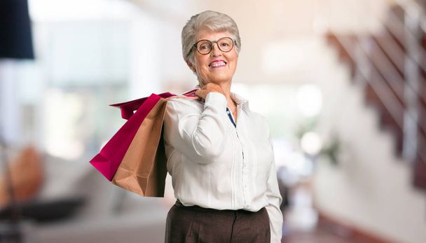 Mooie senior vrouw vrolijk en glimlachend, erg enthousiast uitvoering een shopping tassen, klaar om te gaan winkelen en op zoek naar nieuwe aanbiedingen thuis. - Foto, afbeelding
