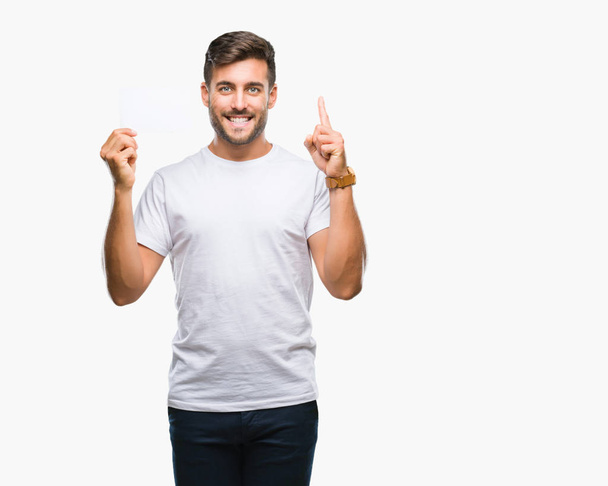Young handsome man holding blank card over isolated background surprised with an idea or question pointing finger with happy face, number one - Fotoğraf, Görsel