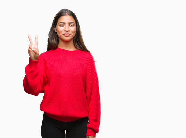 Молодая красивая арабская женщина в зимнем свитере на изолированном фоне показывает и указывает пальцами номер два, улыбаясь уверенно и счастливо
. - Фото, изображение