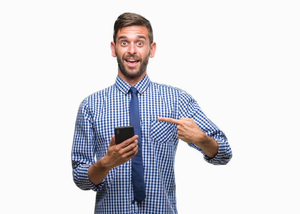 Jonge hadsome man SMS verzenden bericht via smartphone over geïsoleerde achtergrond erg blij met de hand en de vinger te wijzen - Foto, afbeelding