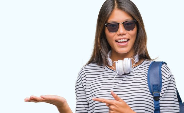 Young asian student woman wearing headphones and backpack over isolated background amazed and smiling to the camera while presenting with hand and pointing with finger. - Φωτογραφία, εικόνα