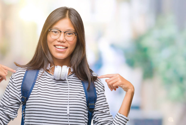 Jeune étudiante asiatique femme portant casque et sac à dos sur fond isolé regardant confiant avec sourire sur le visage, se pointant avec les doigts fiers et heureux
. - Photo, image