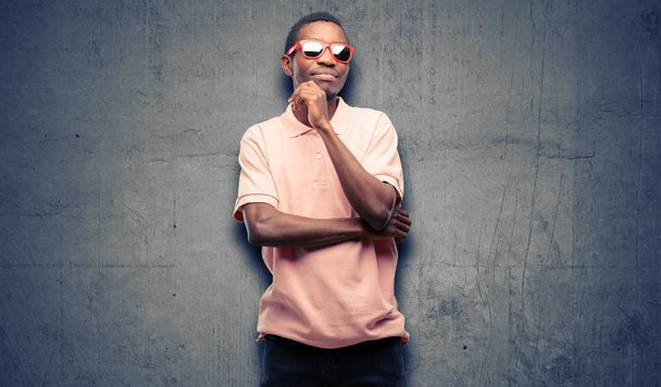 Afrikanischer schwarzer Mann mit Sonnenbrille denkt nach und blickt auf und äußert Zweifel und Verwunderung - Foto, Bild