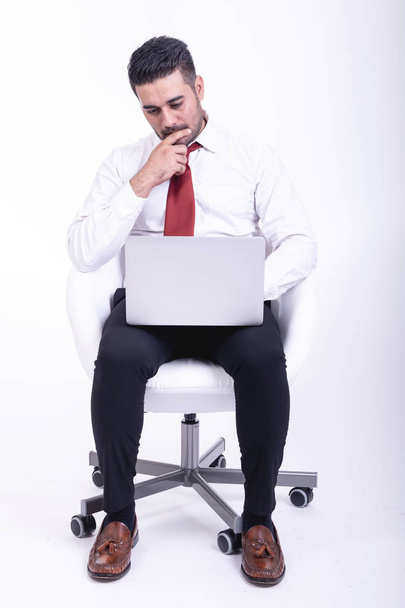 Businessman sitting on white chair isolated. Handsome young indian businessman using laptop portrait, thinking looks. Full length shot. - Foto, afbeelding