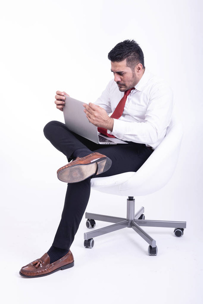 Zakenman zittend op witte stoel geïsoleerd. Knappe jonge Indiase zakenman met laptop portret, erg boos kijkt. Volledige lengte kant schoot. - Foto, afbeelding