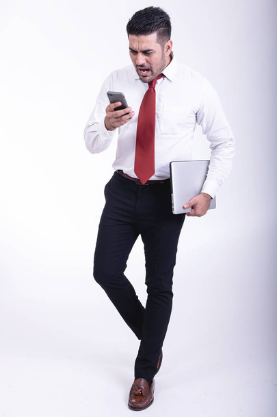 Businessman isolated. Handsome young indian businessman holding laptop, using a phone talking, very upset looks. Full length shot. - Fotografie, Obrázek