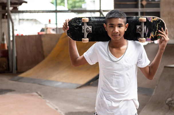 Skateboarder portrait. Smiling Asian boy in urban skateboard park under the bridge standing on floor holding his board. Natural light setting. - Photo, image