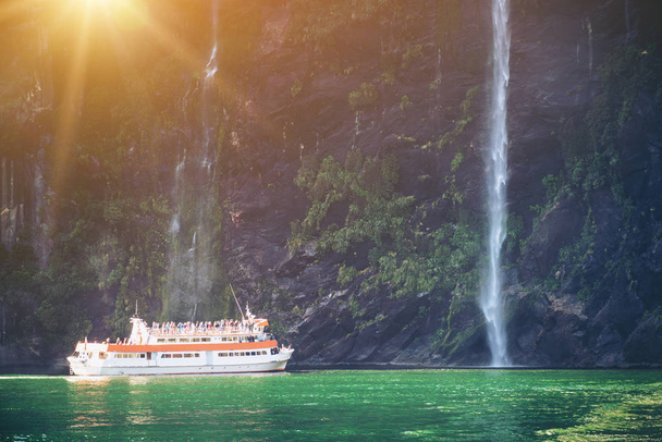 Ausflugsboot mit Touristen nähert sich großem Wasserfall im Milford Sound. schöne landschaftlich reizvolle Kreuzfahrt durch den Fiordland-Nationalpark auf der Südinsel Neuseelands. - Foto, Bild