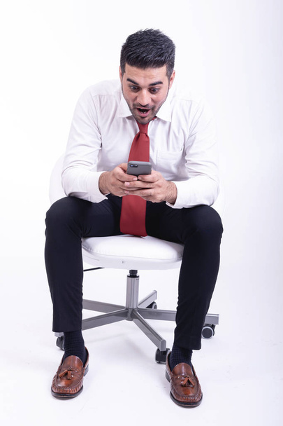 Businessman sitting on white chair isolated. Handsome young indian businessman using mobile phone, shock looks.  Full length shot. - Fotografie, Obrázek
