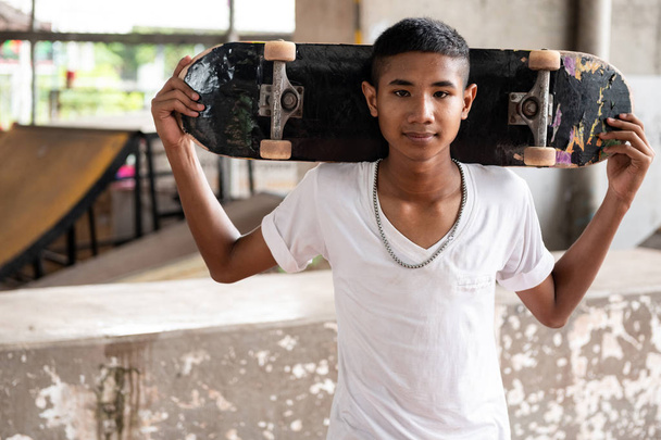 Skateboarder portrait. Smiling Asian boy in urban skateboard park under the bridge standing on floor holding his board. Natural light setting. - 写真・画像