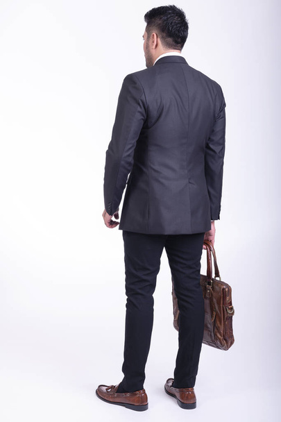 Businessman isolated. Handsome young indian businessman in suit with bag portrait, hands in pocket smile looks. Full length back shot. - Fotó, kép