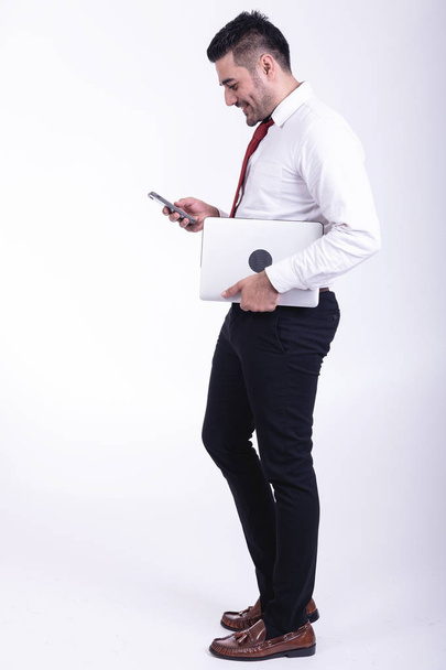 Businessman isolated. Handsome young indian businessman holding laptop, looking at his phone, confident looks. Full length side shot. - Zdjęcie, obraz