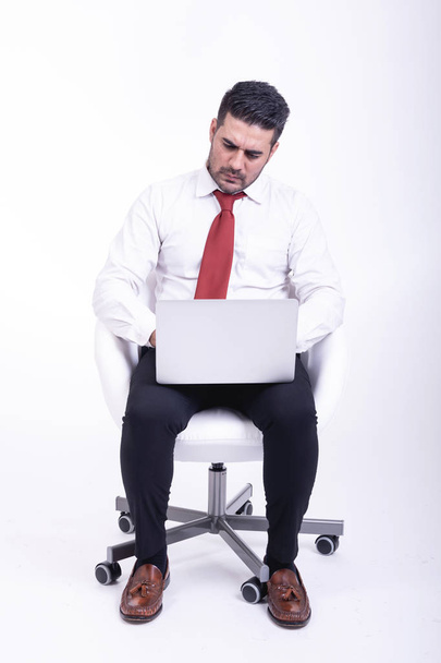 Businessman sitting on white chair isolated. Handsome young indian businessman using laptop portrait, upset looks. Full length side shot. - Foto, imagen
