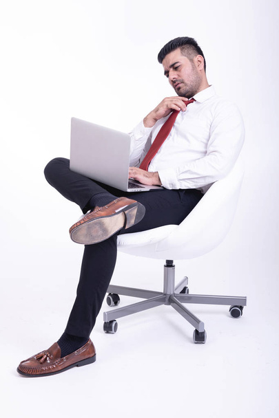 Businessman sitting on white chair isolated. Handsome young indian businessman using laptop portrait, thinking looks. Full length side shot. - Fotó, kép