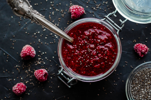 Homemade raspberry jam with chia seeds in a jar with a spoon inside on black background. Healthy idea for breakfast toasts. Superfoods and healthy food concept. Top view. - Photo, Image