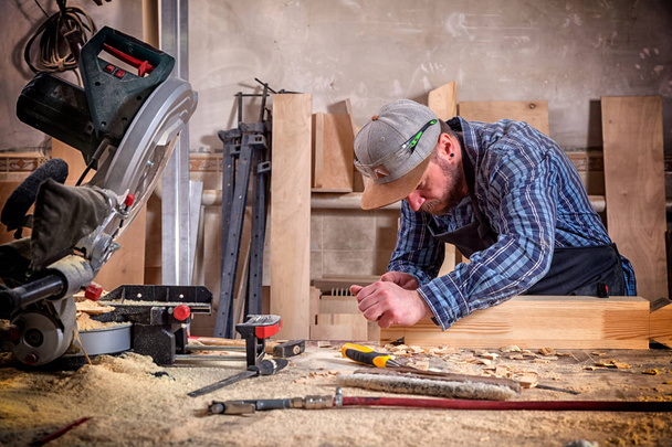 опытный плотник в рабочей одежде и мелкий предприниматель измеряет деревянную доску линейкой и отмечает карандашом необходимые точки для ломтиков
 - Фото, изображение