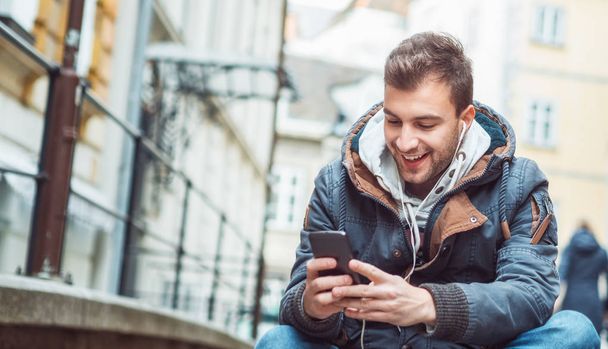 Szczęśliwy młody człowiek trzyma jego smartphone podczas pisania wiadomości SMS, coś i siedzi na ulicy  - Zdjęcie, obraz